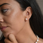 Side profile of a model wearing a large ivory pearl button stud earring, with her hand resting beneath her chin.